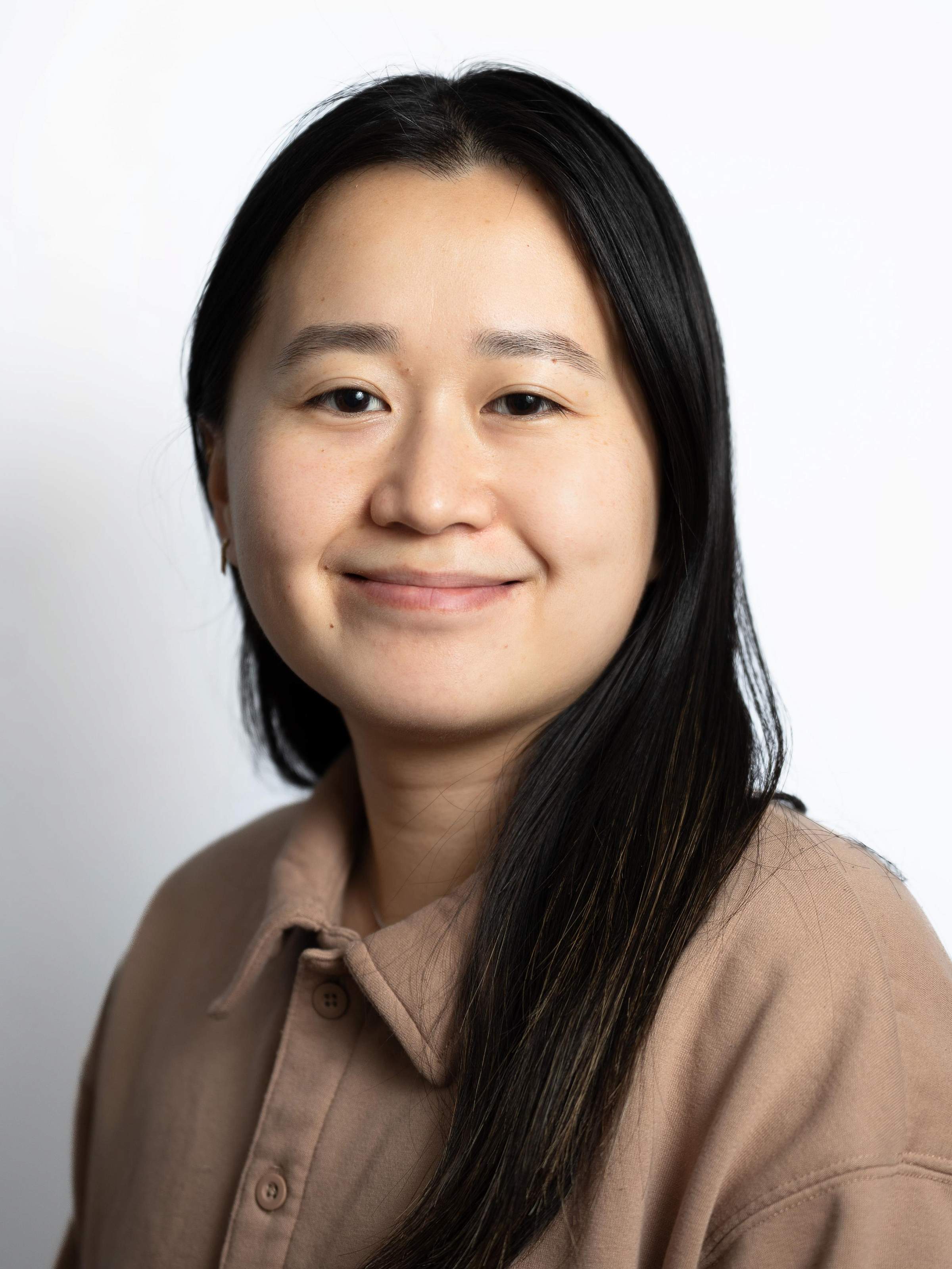 A portrait of Shiya, a Chinese person with long black hair and wearing a brown collared shirt, looking into the camera and smiling. Posing in front of a blank white background.